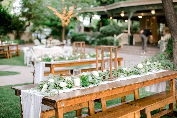 A rustic wooden farm table and bench set decorated with a white runner, greenery, and white flowers at an outdoor event.