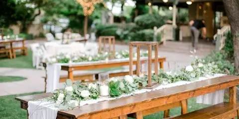 A rustic wooden farm table and bench set decorated with a white runner, greenery, and white flowers at an outdoor event.