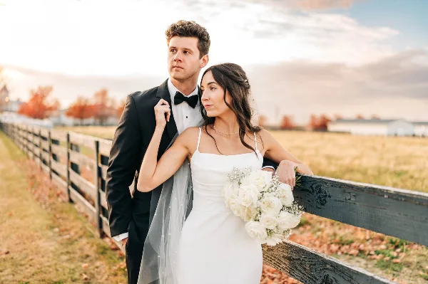 A bride and groom posing together outdoors near a wooden fence, with the bride holding a bouquet of white roses.