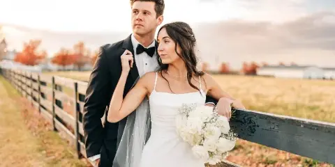 A bride and groom posing together outdoors near a wooden fence, with the bride holding a bouquet of white roses.