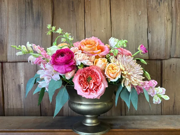 A lush floral arrangement featuring pink and peach roses, ranunculus, and alstroemeria in a brass urn-style vase against a rustic wooden background.