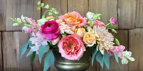 A lush floral arrangement featuring pink and peach roses, ranunculus, and alstroemeria in a brass urn-style vase against a rustic wooden background.
