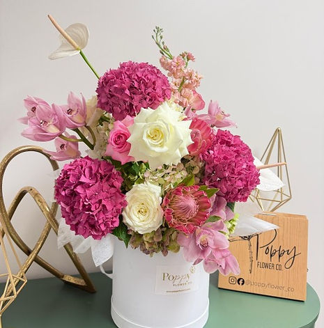 A floral arrangement with pink and white flowers in a white bucket, with a Poppy Flower Co. sign in the background.