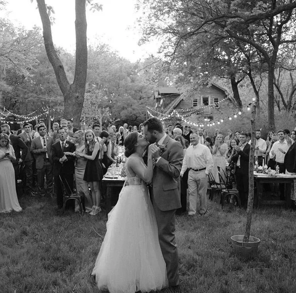 Black and white photo of a bride and groom kissing at an outdoor wedding reception with guests, long tables, string lights, and a rustic building in a wooded setting.