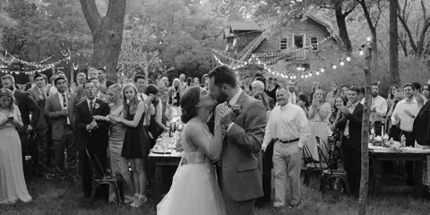 Black and white photo of a bride and groom kissing at an outdoor wedding reception with guests, long tables, string lights, and a rustic building in a wooded setting.