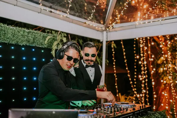 A DJ wearing headphones and sunglasses operates a mixing console at an event venue decorated with string lights.