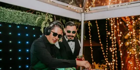 A DJ wearing headphones and sunglasses operates a mixing console at an event venue decorated with string lights.