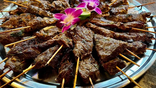 A silver platter filled with grilled beef satay skewers garnished with a purple orchid and lime slice.