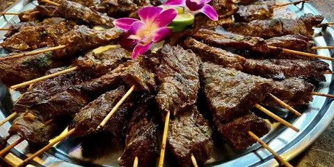 A silver platter filled with grilled beef satay skewers garnished with a purple orchid and lime slice.
