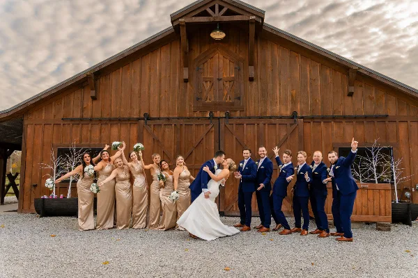 A wedding party posing in front of a large wooden barn structure.