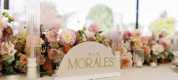 A personalized lettered wedding sign reading 'MORALES' displayed on a table with floral arrangements and tall glass candle holders.
