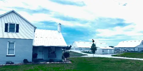 Rustic farm wedding venue with a white farmhouse in the foreground and multiple barns in the background under a cloudy sky.