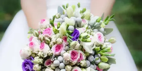 A close-up of a bride holding a large, round wedding bouquet featuring pink roses, white calla lilies, and greenery.