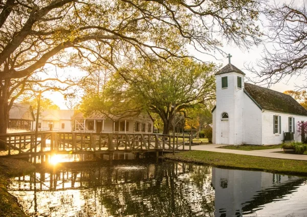 A historic white church with a cross, a wooden bridge, and traditional houses in a village setting at sunset.