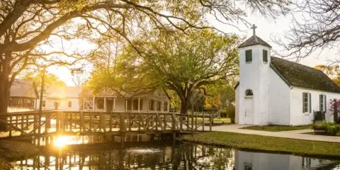A historic white church with a cross, a wooden bridge, and traditional houses in a village setting at sunset.