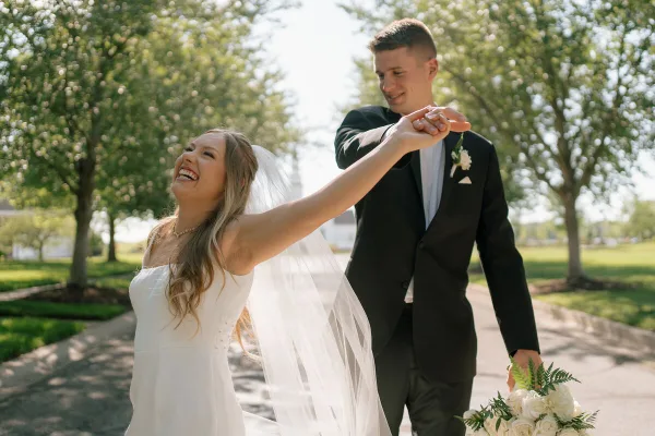 A bride and groom laughing and dancing outdoors on a sunny day.