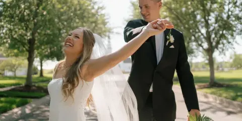 A bride and groom laughing and dancing outdoors on a sunny day.