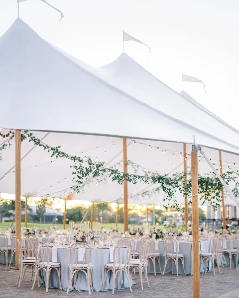 A white wedding tent interior with round tables, floral centerpieces, and hanging greenery.