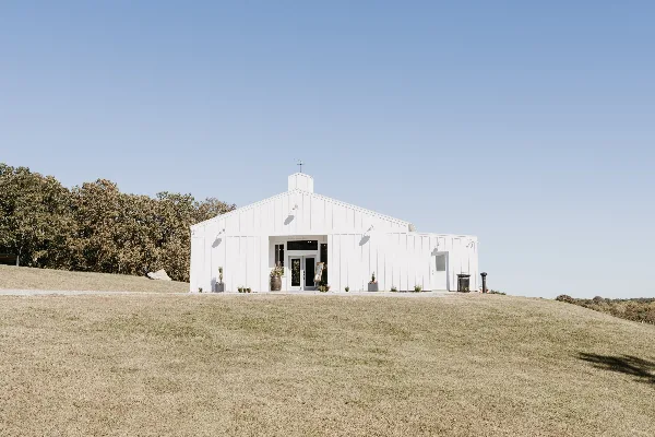 A white barn-style building with a gabled roof and cupola sits on a grassy hill under a clear blue sky.
