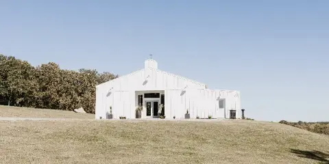 A white barn-style building with a gabled roof and cupola sits on a grassy hill under a clear blue sky.
