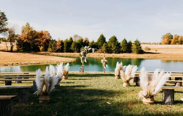Outdoor wedding ceremony setup with a wooden arch and pampas grass decorations on a lawn next to a pond.