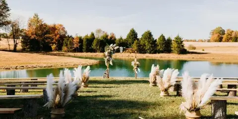 Outdoor wedding ceremony setup with a wooden arch and pampas grass decorations on a lawn next to a pond.