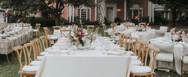 An outdoor wedding reception setup with white tablecloths, floral centerpieces, and wooden chairs with cushions.