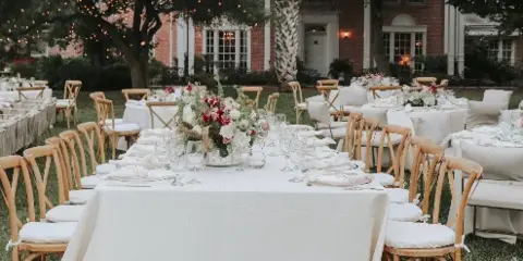 An outdoor wedding reception setup with white tablecloths, floral centerpieces, and wooden chairs with cushions.