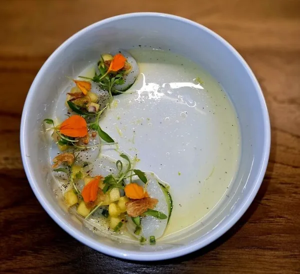 Plated Hokkaido scallop ceviche with edible flowers, cucumber, and tropical salsa in a white bowl on a wooden table.