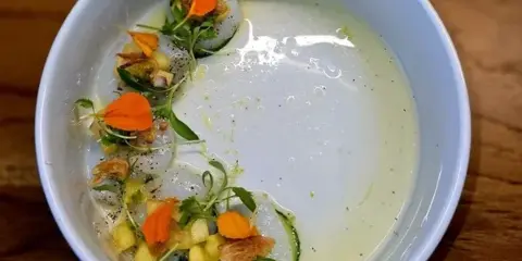 Plated Hokkaido scallop ceviche with edible flowers, cucumber, and tropical salsa in a white bowl on a wooden table.
