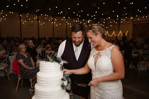 A bride and groom cutting a wedding cake in a large indoor venue with string lights and guests seated at tables.