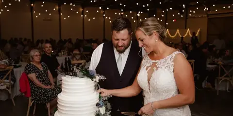 A bride and groom cutting a wedding cake in a large indoor venue with string lights and guests seated at tables.
