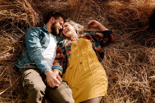 A couple lying in dry grass, smiling and holding hands, wearing casual outdoor clothing.