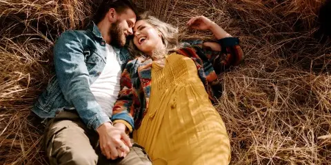 A couple lying in dry grass, smiling and holding hands, wearing casual outdoor clothing.