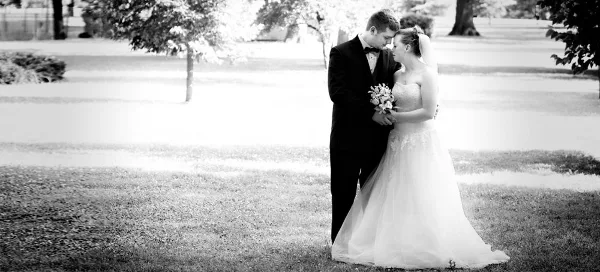 A black-and-white photograph of a bride and groom standing together outdoors, with the groom in a tuxedo and the bride in a wedding gown holding a bouquet.