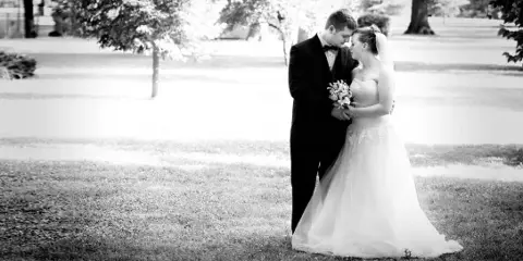 A black-and-white photograph of a bride and groom standing together outdoors, with the groom in a tuxedo and the bride in a wedding gown holding a bouquet.