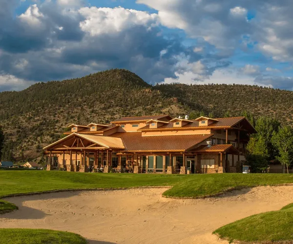 Exterior view of a large mountain resort clubhouse with a golf course green and sand bunker in the foreground, set against forested hills.