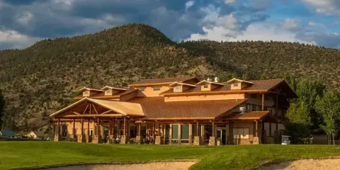 Exterior view of a large mountain resort clubhouse with a golf course green and sand bunker in the foreground, set against forested hills.