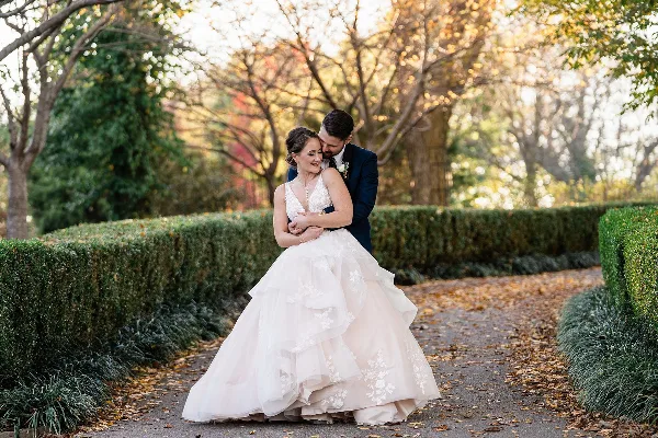 A bride in a white wedding gown with styled hair and makeup embracing a groom in a garden setting.