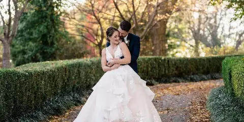 A bride in a white wedding gown with styled hair and makeup embracing a groom in a garden setting.