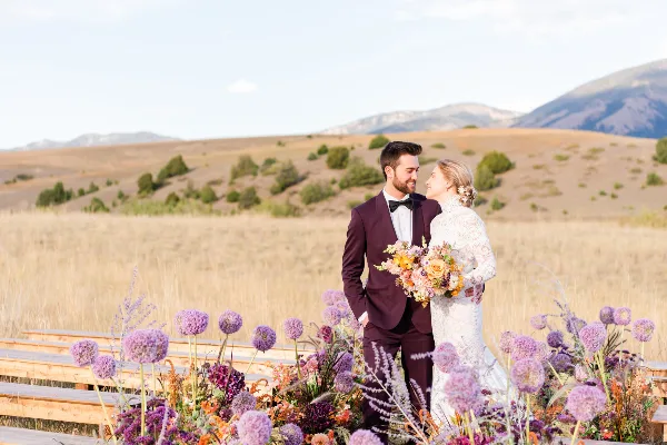 A bride and groom standing together in a field with mountains in the background, holding a bouquet.