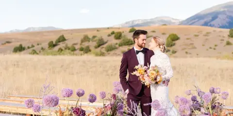 A bride and groom standing together in a field with mountains in the background, holding a bouquet.