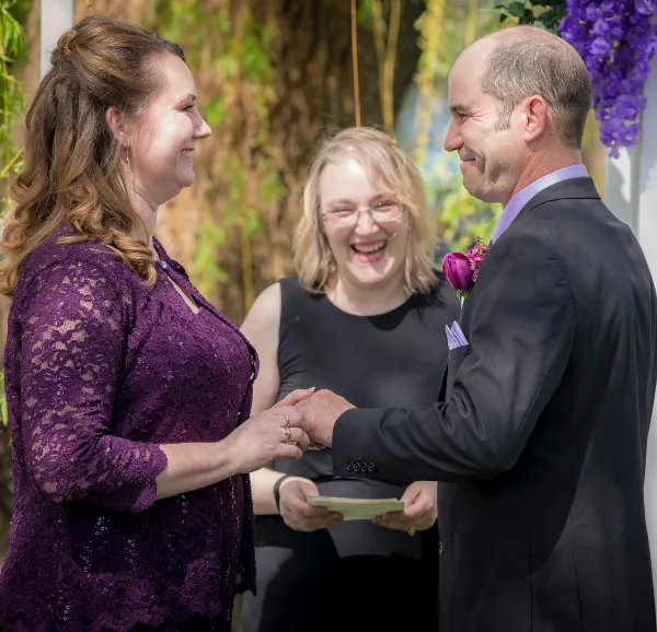 A bride and groom exchanging rings during an outdoor wedding ceremony officiated by a woman.