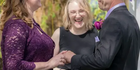 A bride and groom exchanging rings during an outdoor wedding ceremony officiated by a woman.