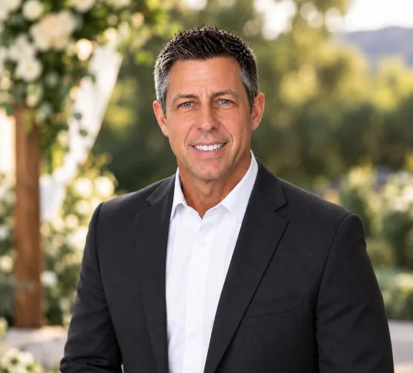A smiling man in a black suit and white shirt standing in front of a wedding floral arch.