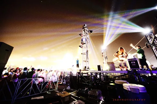 A DJ performing on an outdoor stage at night with bright stage lights, a large crowd, and visible DJ equipment.