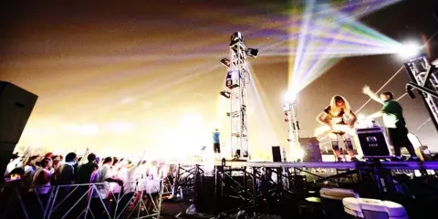 A DJ performing on an outdoor stage at night with bright stage lights, a large crowd, and visible DJ equipment.
