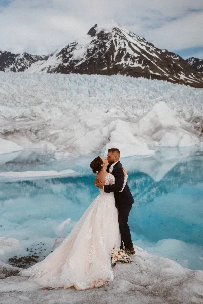 A couple in wedding attire embracing and kissing in front of a blue glacial lake and snow-capped mountains.