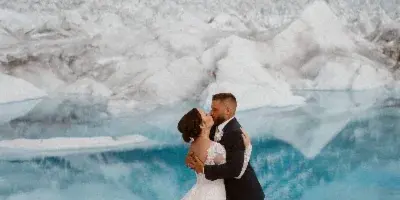 A couple in wedding attire embracing and kissing in front of a blue glacial lake and snow-capped mountains.