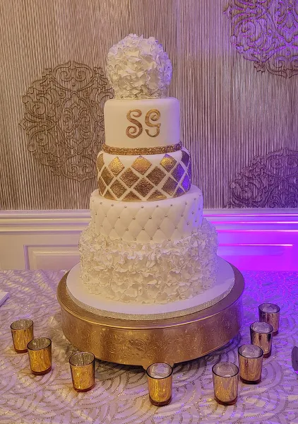 A four-tier white wedding cake featuring gold lattice patterns, pearl accents, and a floral topper, displayed on a gold stand.